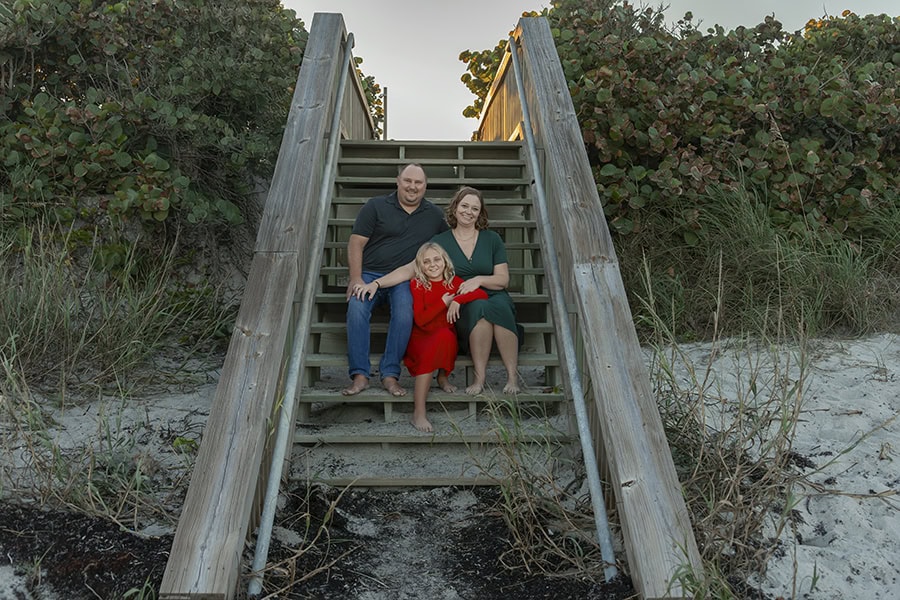 Family of 3 sitting on wooden stairs at the beach.
