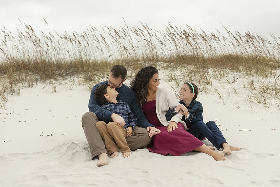 Family of 4 sitting on a beach dune looking at one another.
